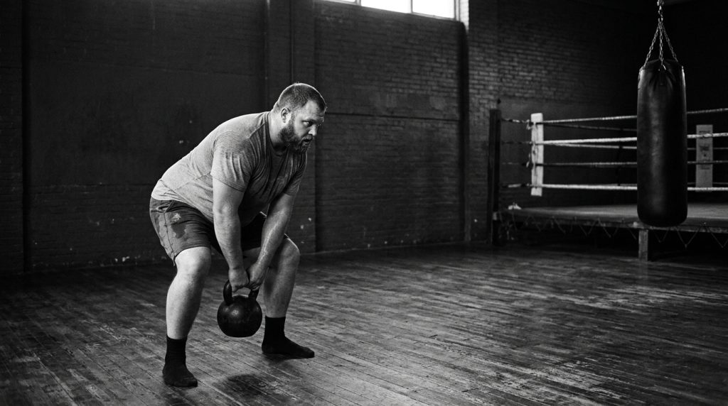 Heavyset man in a grey shirt hinging into a kettlebell swing on a worn wooden gym floor beside an empty ring